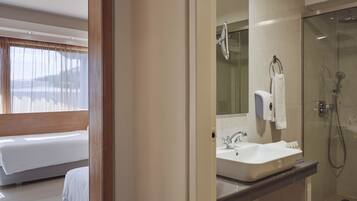 Un baño de hotel moderno con cabina de ducha de vidrio, un lavamanos con espejo y una cama en la habitación contigua.