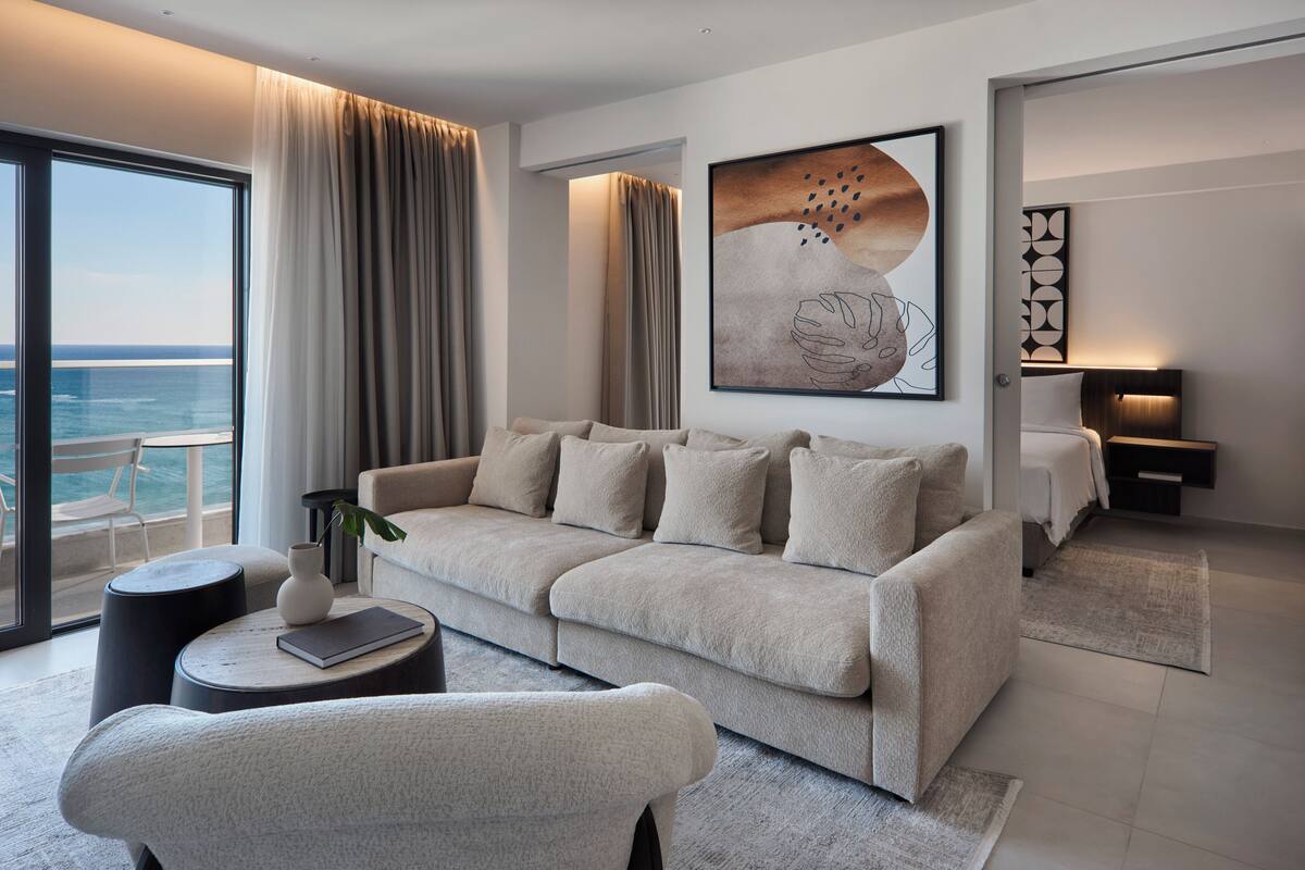 A modern hotel room with a large window overlooking the sea, a sofa, a chair, and a bedside table with a lamp.