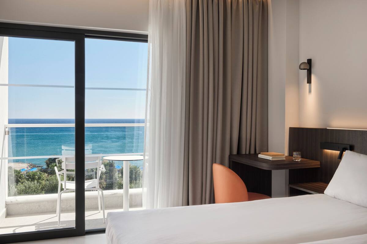 A hotel room with a bed, a desk with a chair, a view of the ocean, and a balcony with a table and chairs.