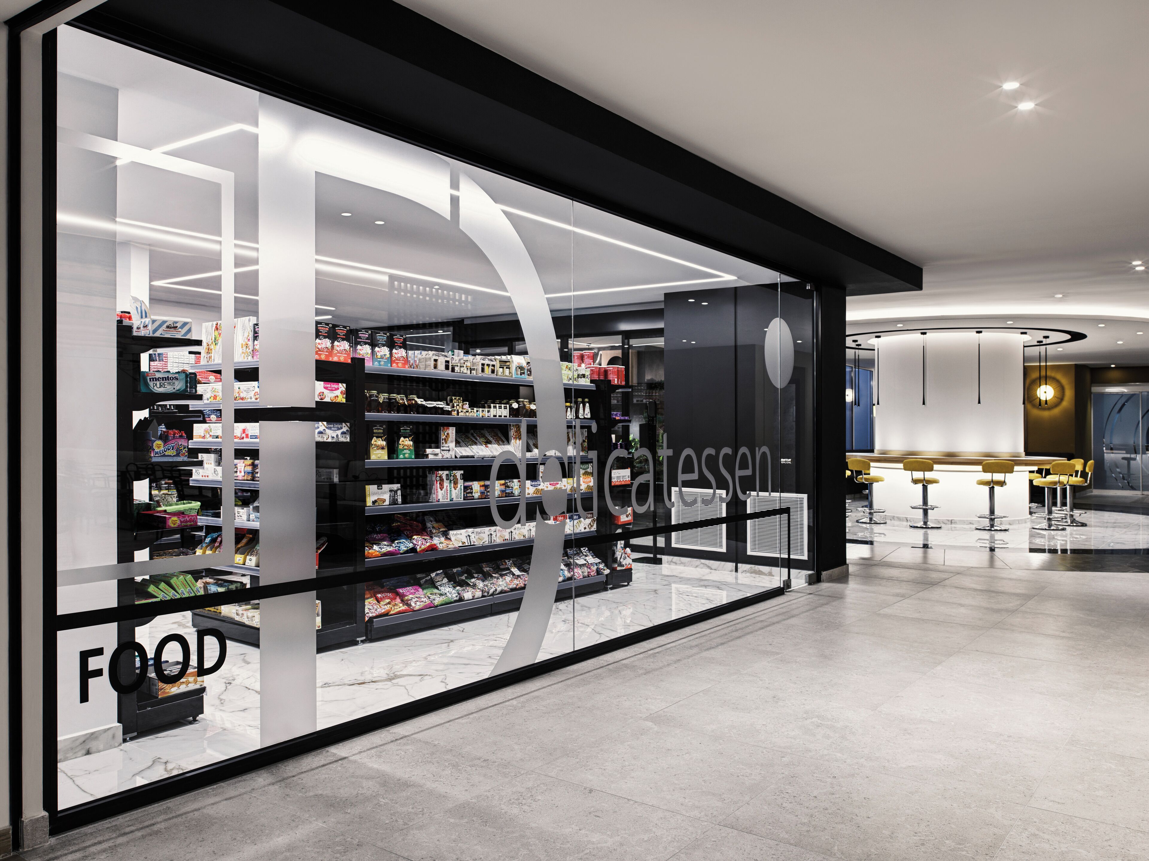 A modern food store with a glass facade, shelves stocked with various products, and a seating area with yellow chairs.