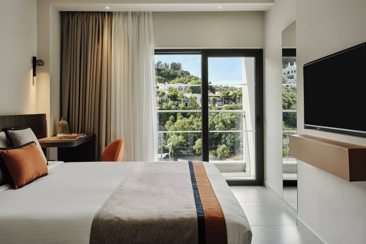 A hotel room with a large bed, a desk with a chair, a TV mounted on the wall, and a view of a cityscape through the window.