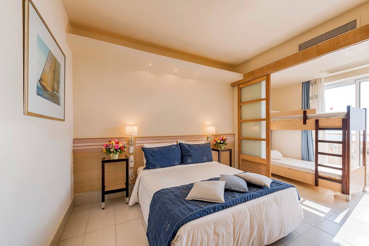 A hotel room with a large bed, a bunk bed, a bedside table with a lamp, a wall-mounted phone, and a window with curtains.