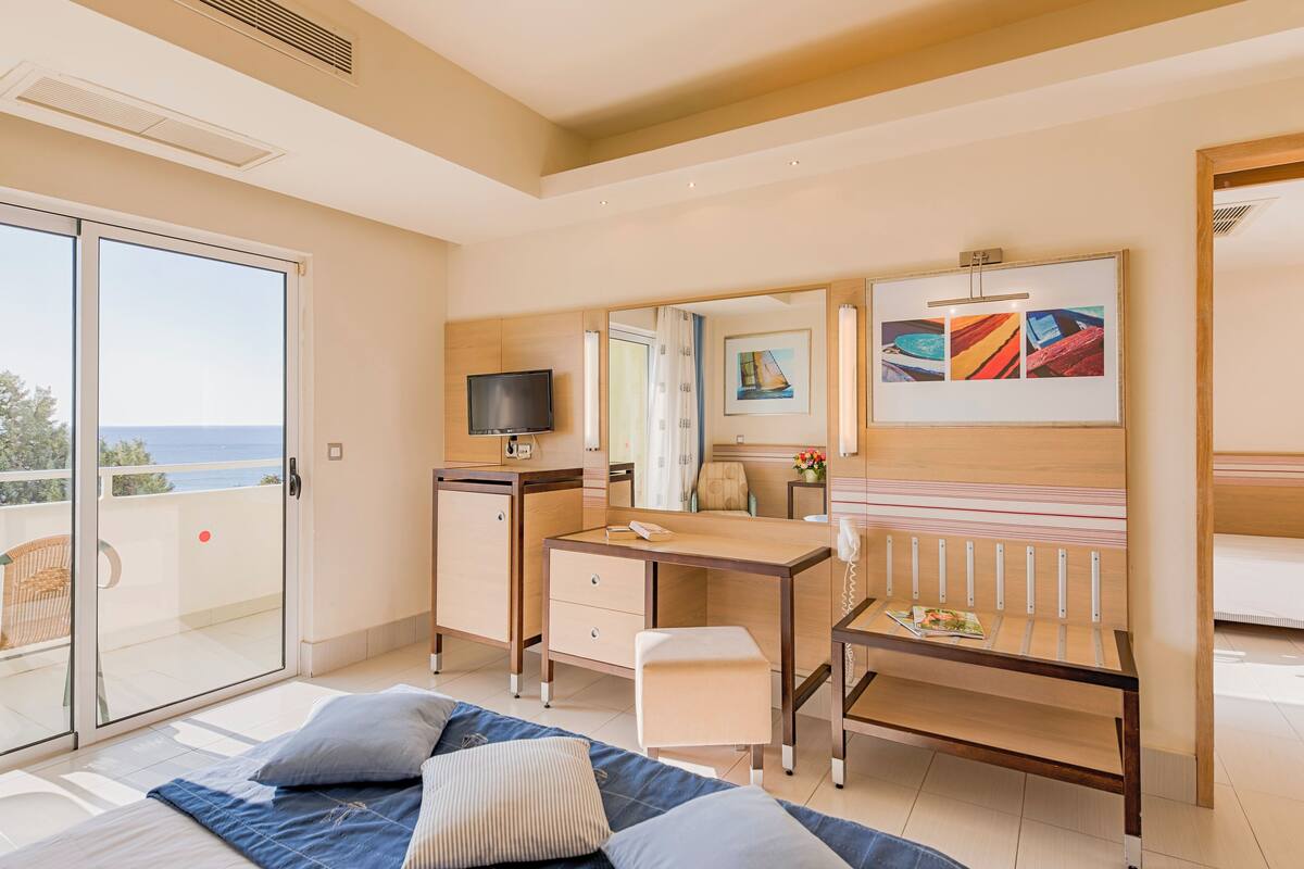 A hotel room with a bed, a desk, a chair, a TV, and a balcony with a view of the sea.