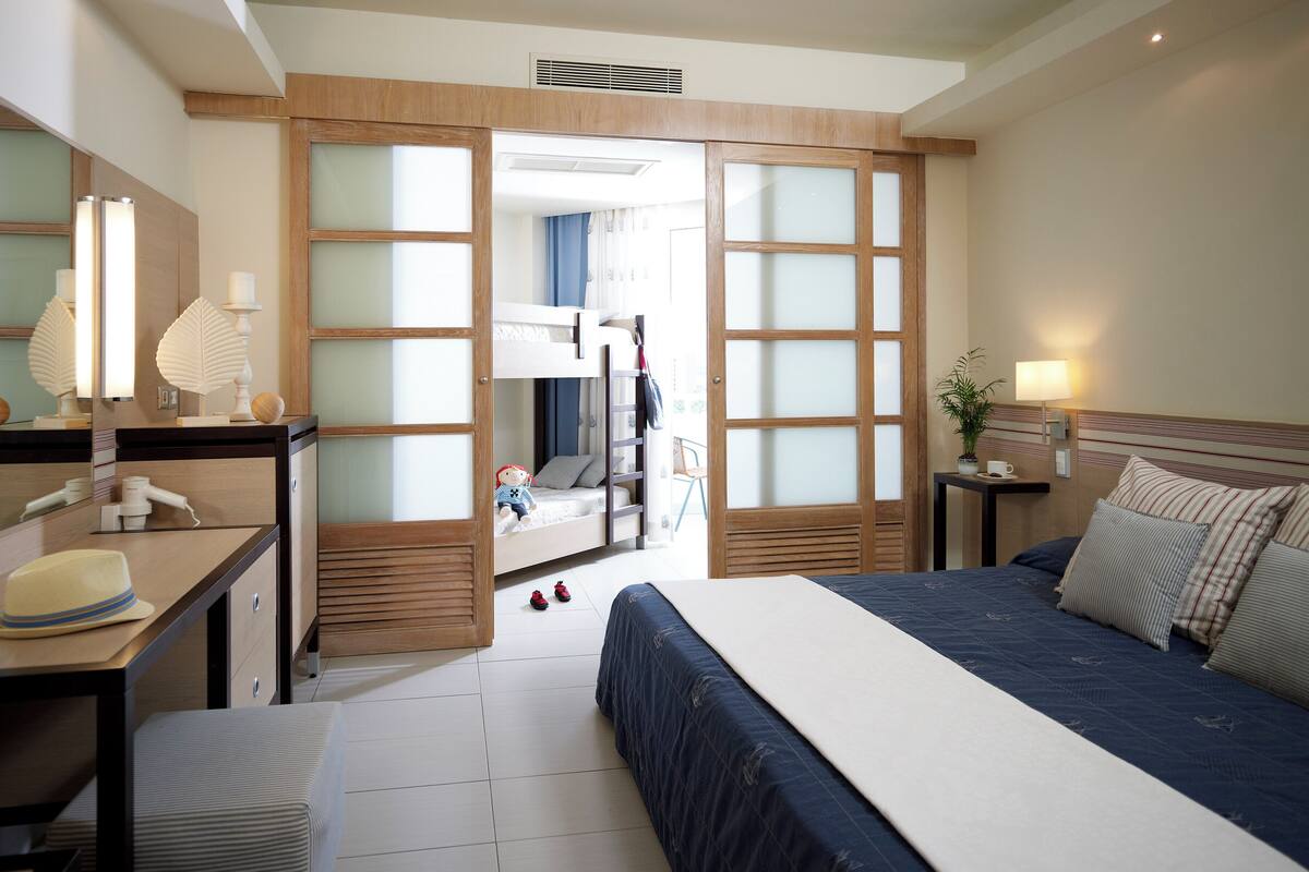 A hotel room with a bunk bed, a desk with a lamp, a bedside table with a plant, and a large window.