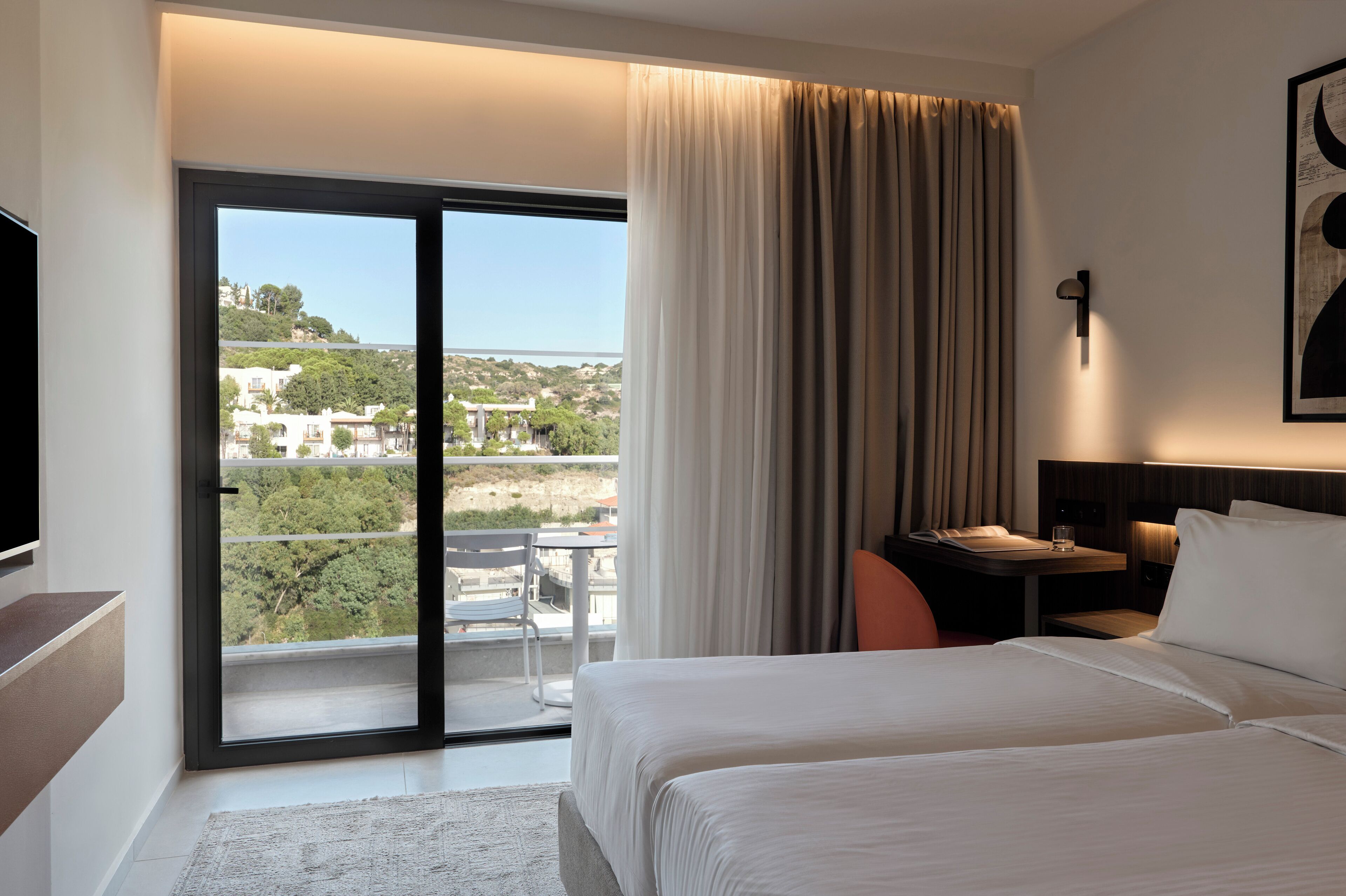Hotelkamer met een bed, een bureau, een stoel en uitzicht op de stad door een groot raam.