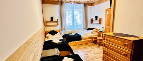 A small, cozy room with two single beds, a wooden dresser, a desk, and a window with curtains.