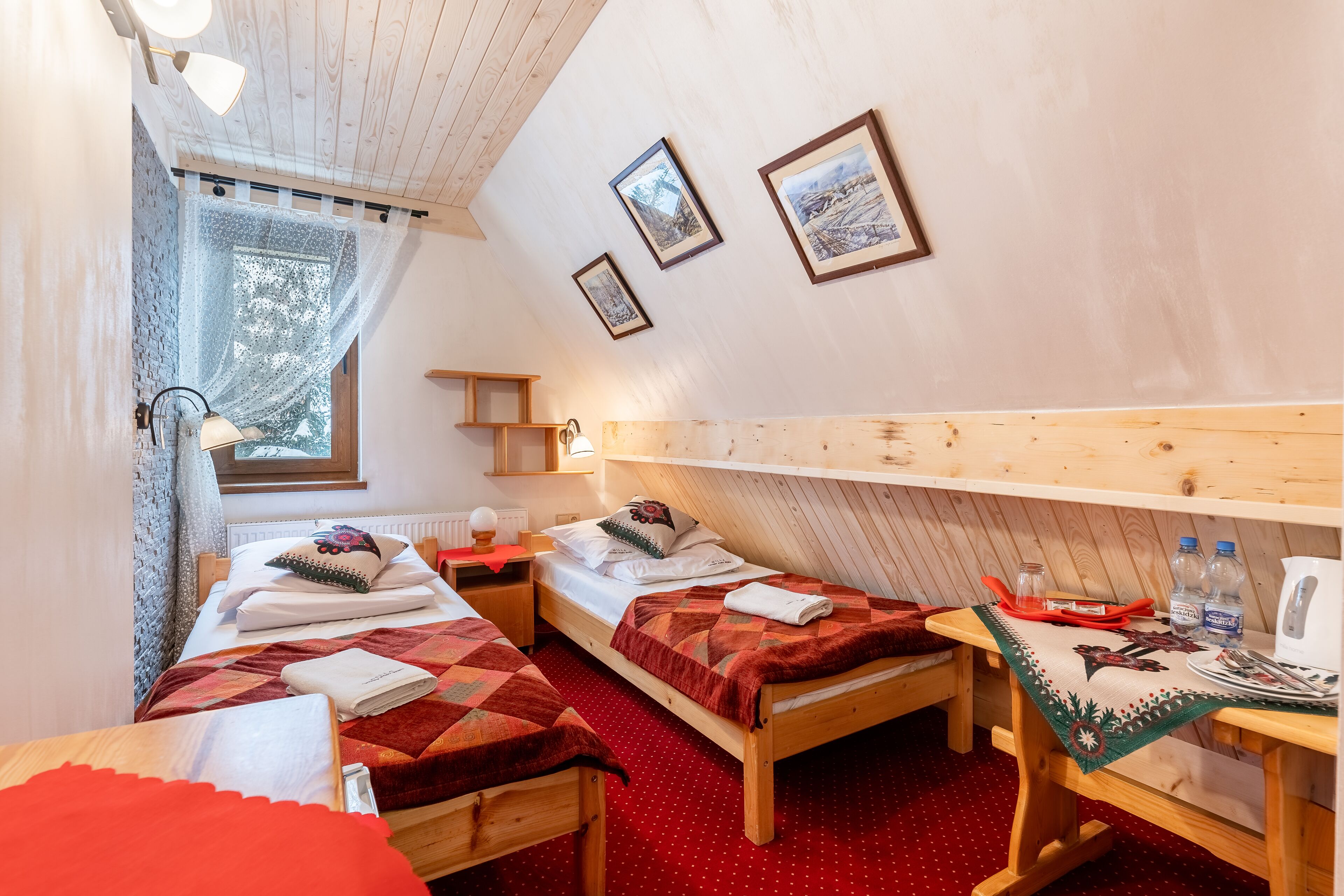 A cozy room with two wooden beds, a small table with a kettle, and framed pictures on the wall.