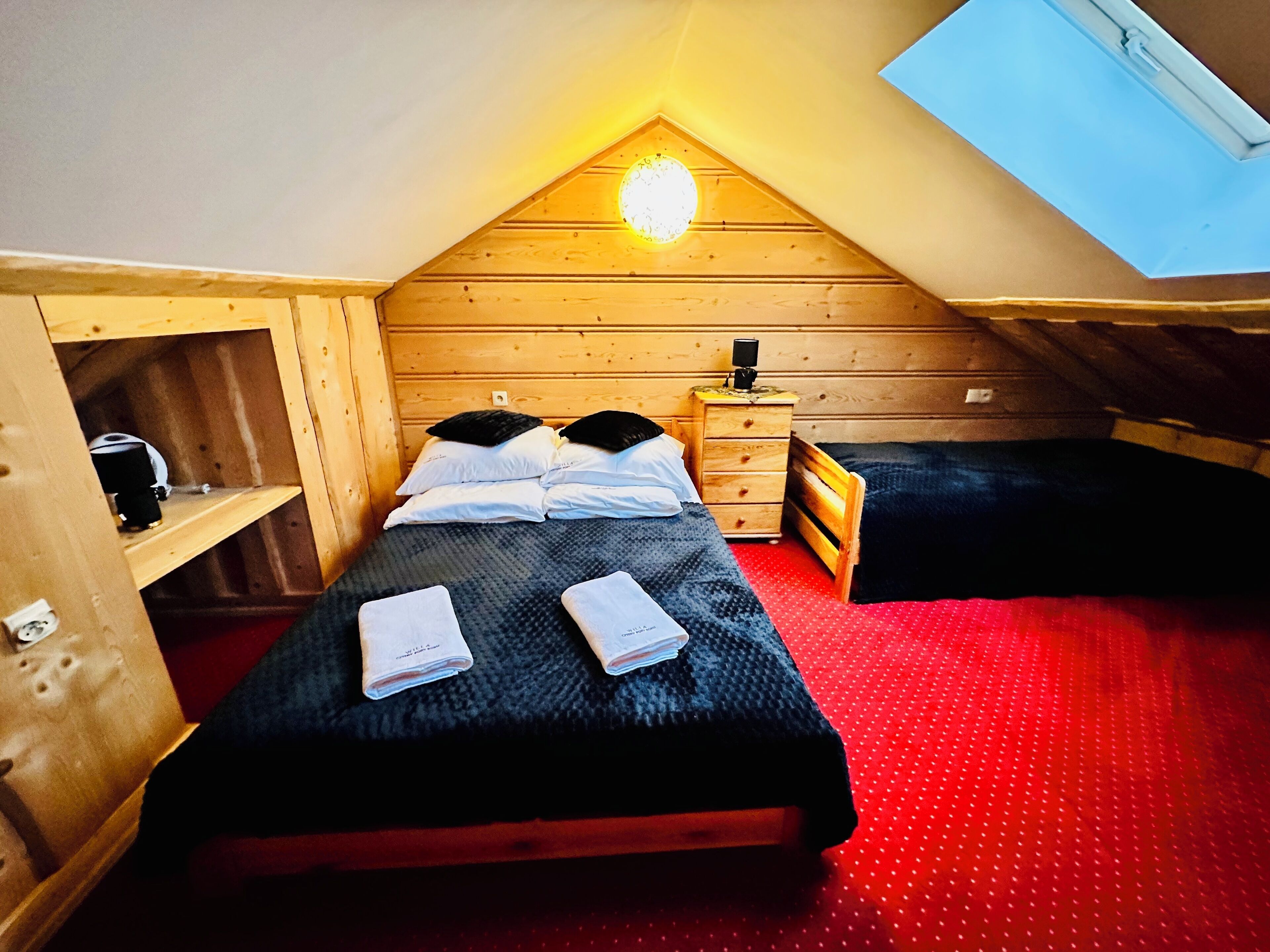A small, cozy attic bedroom with a sloped ceiling, a skylight, a wooden dresser, a bedside table, and a single bed with pillows and a blanket.