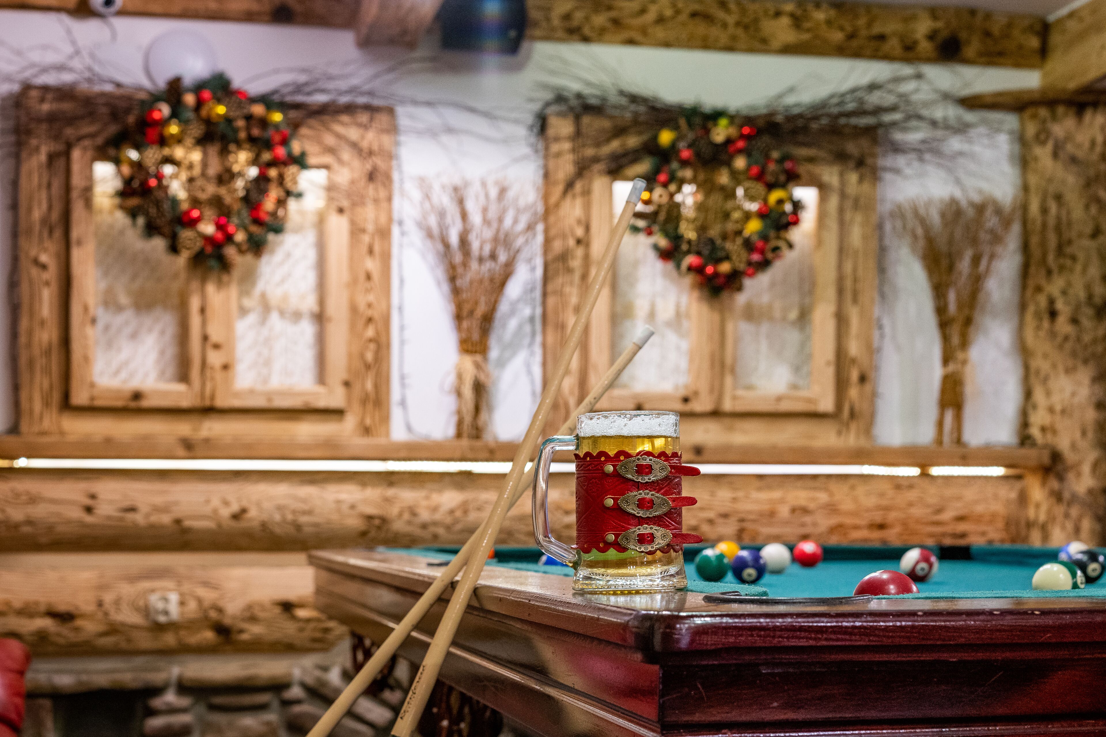 A pool table with a beer mug and pool cues, surrounded by Christmas wreaths and a rustic wooden wall.