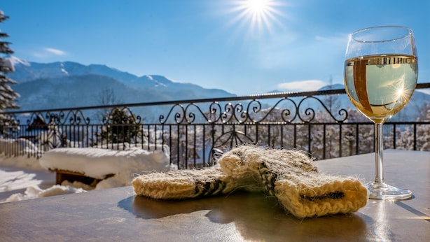 Une table avec un verre de vin blanc et une paire de moufles, le tout placé devant un paysage de montagne enneigé.