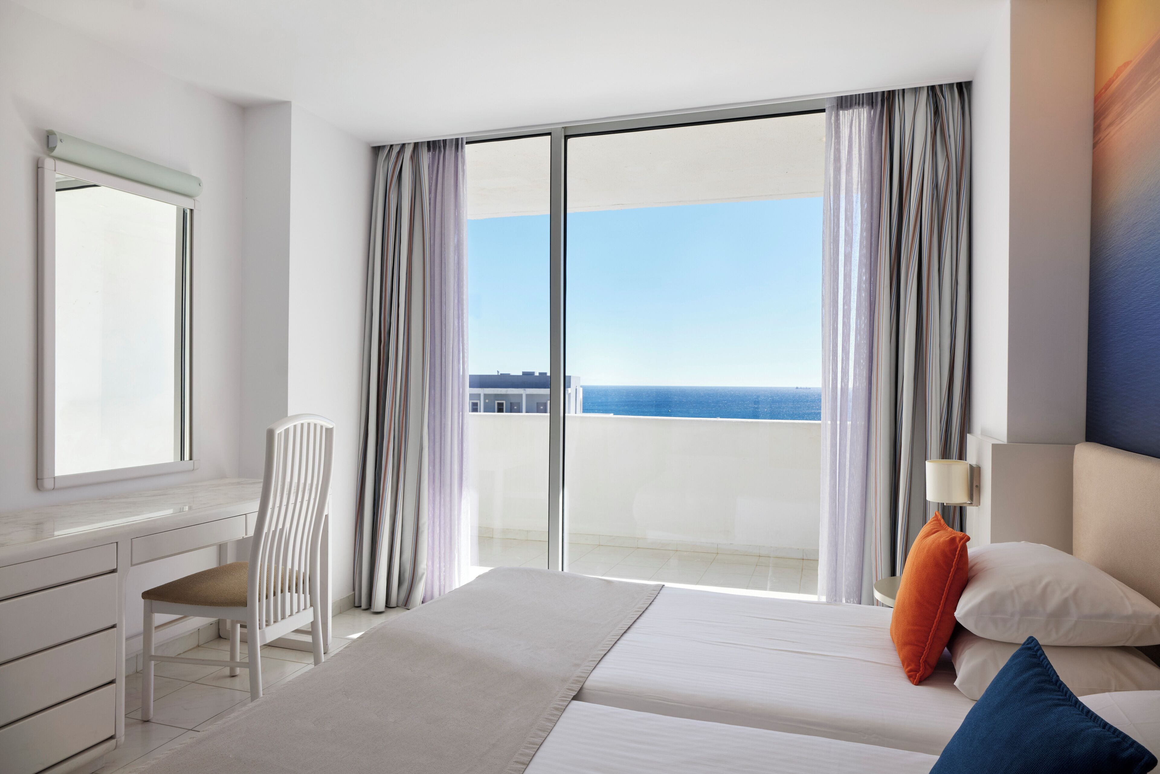 A hotel room with a bed, a desk, a chair, and a view of the sea.