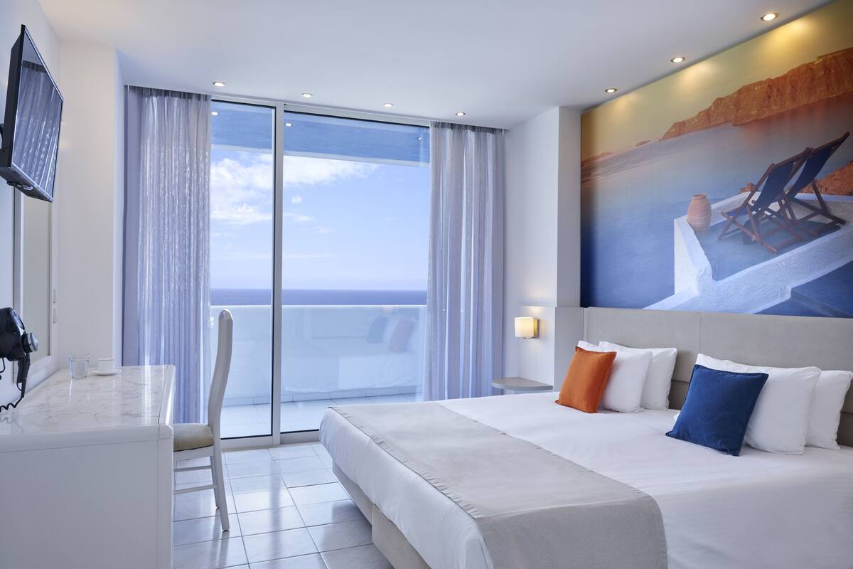 A hotel room with a large bed, a balcony with a view, a desk with a chair, and a TV.