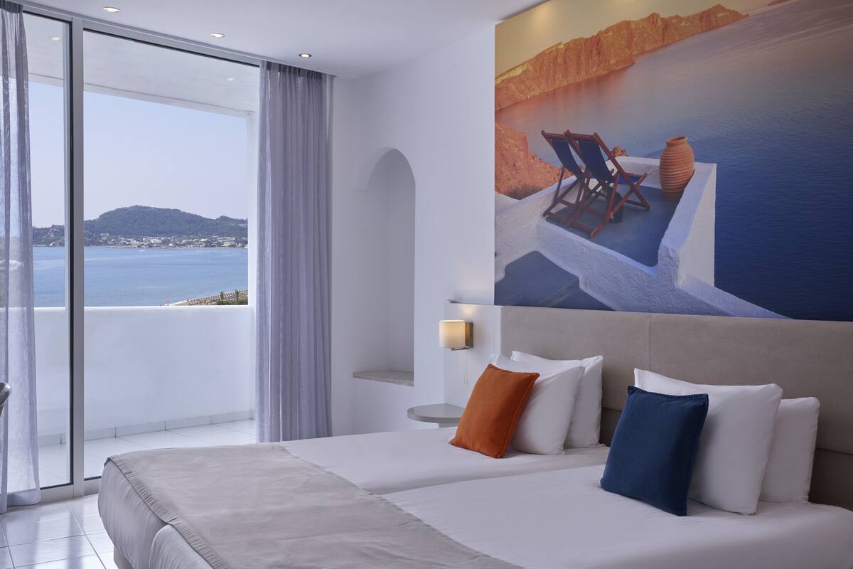 A hotel room with a large bed, a nightstand, a wall-mounted lamp, and a view of the sea.