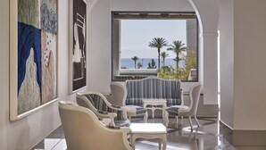 A modern interior with a seating area, a large window with a view of palm trees and the sea, and artwork on the walls. - Epsilon Hotel Apartments (Rhodes)