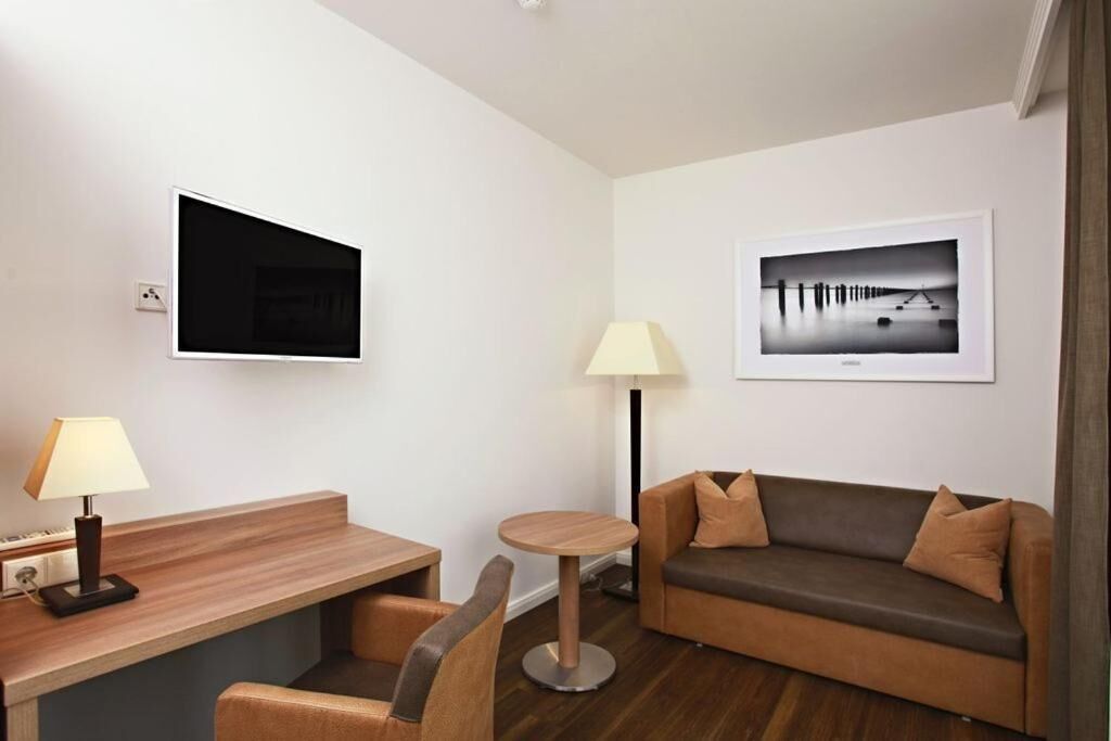 Quarto de hotel moderno com escrivaninha de madeira, mesa redonda, sofá, televisão fixada na parede e duas molduras com imagens.