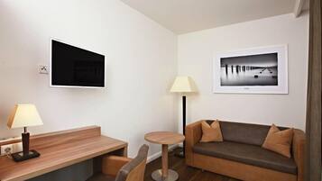 Quarto de hotel moderno com escrivaninha de madeira, mesa redonda, sofá, televisão fixada na parede e duas molduras com imagens.