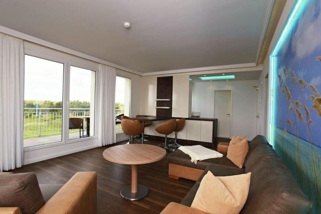 A modern living room with a round wooden table, two brown armchairs, a dark brown sofa, and a large window with curtains.