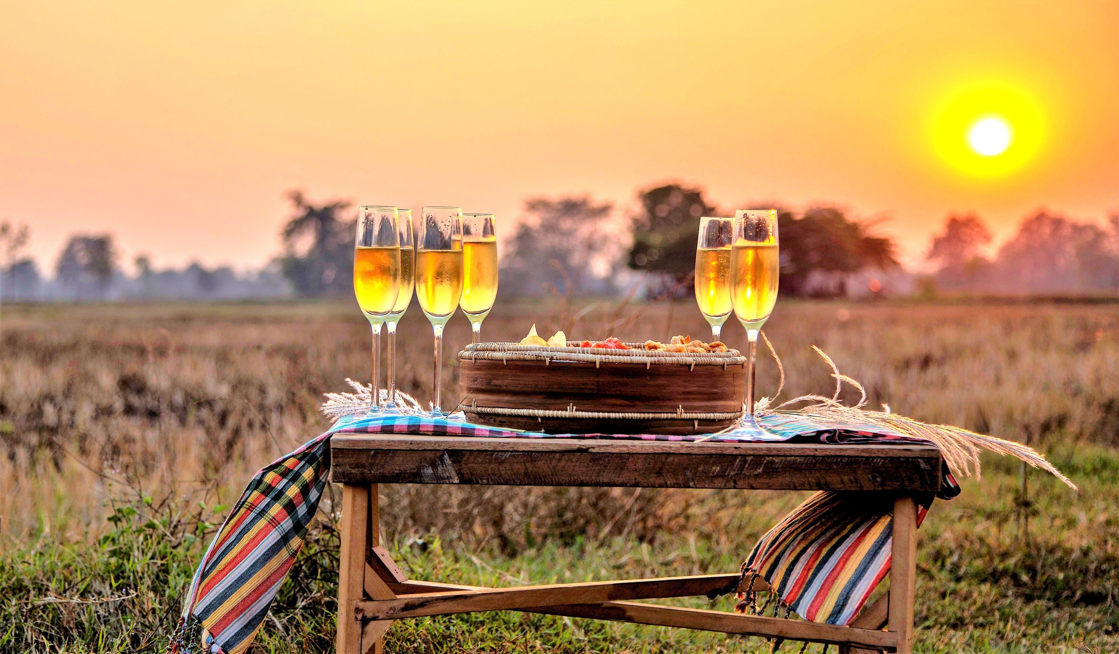 Un montaje para picnic con una mesa de madera, una cesta de aperitivos y cuatro vasos de una bebida amarilla, sobre un fondo de atardecer.
