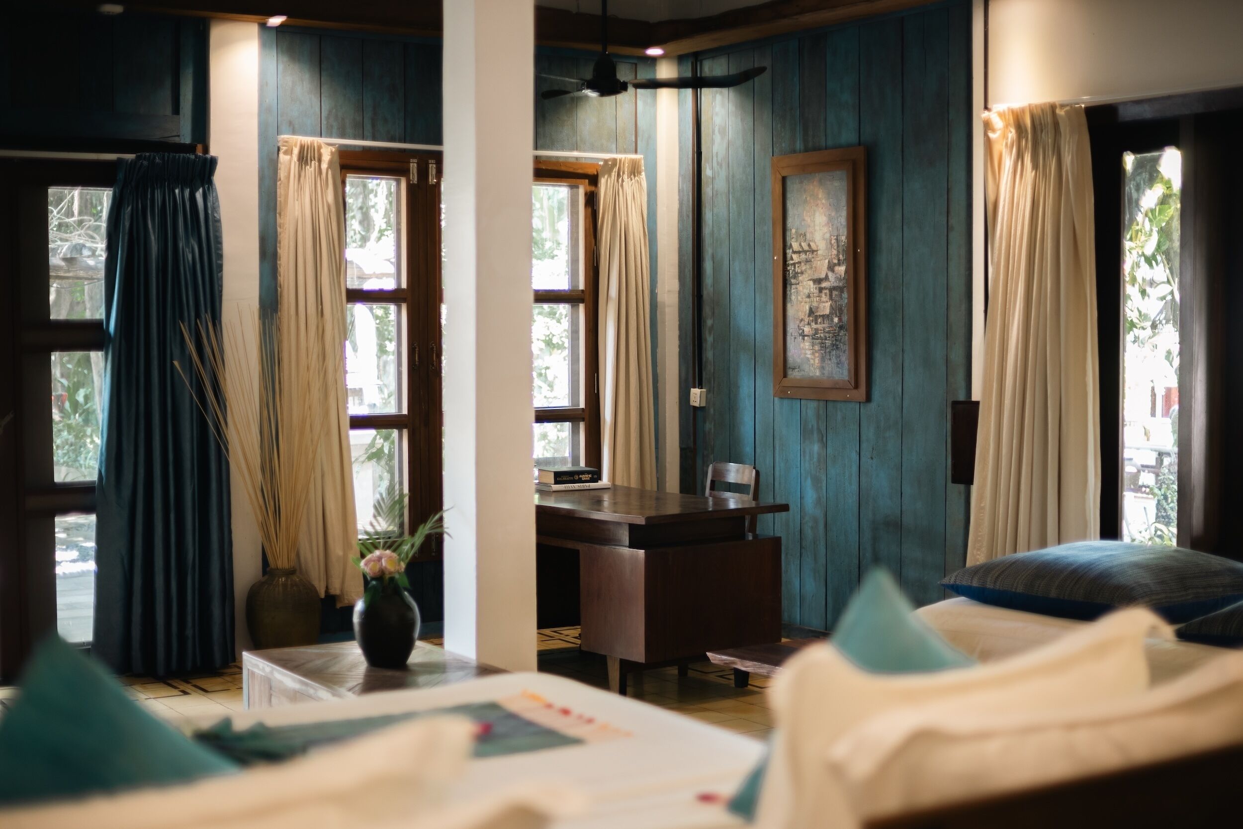 Una habitación con una cama, un escritorio y una silla. Hay cortinas en las ventanas y un jarrón con flores sobre el escritorio.