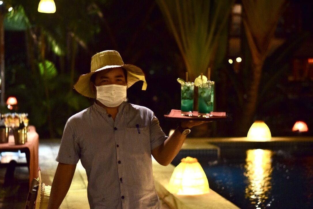 A person holding a tray with two drinks and a bottle, standing by a pool with a fountain and palm trees in the background.