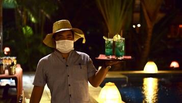 A person holding a tray with two drinks and a bottle, standing by a pool with a fountain and palm trees in the background.