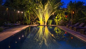 Una piscina nocturna con una fuente central, rodeada de sillones de descanso y plantas tropicales.