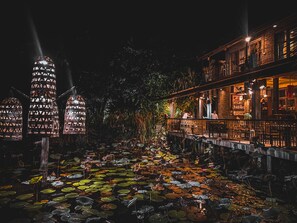 Vista nocturna de un edificio con un área exterior cubierta, que incluye un estanque con lirios y faroles decorativos.