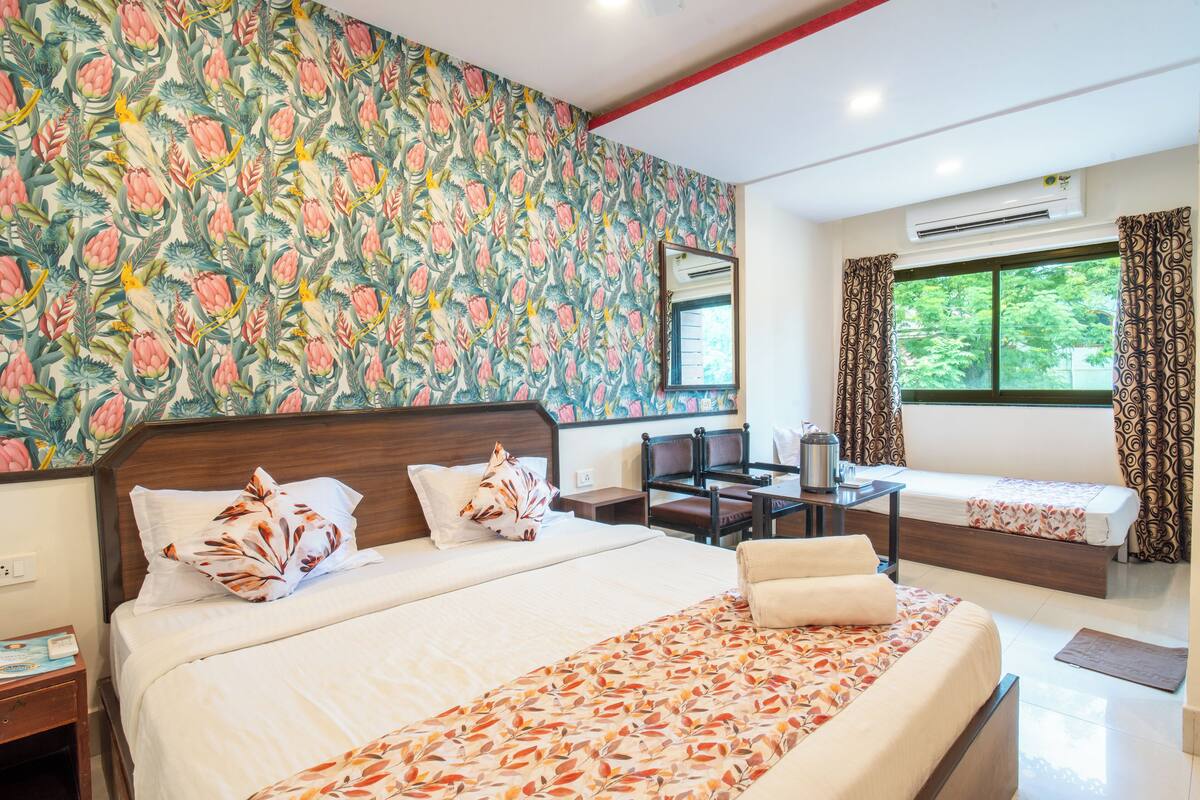 A hotel room with a large bed, a desk, a chair, and a small table. There is a window with curtains and an air conditioning unit above it.