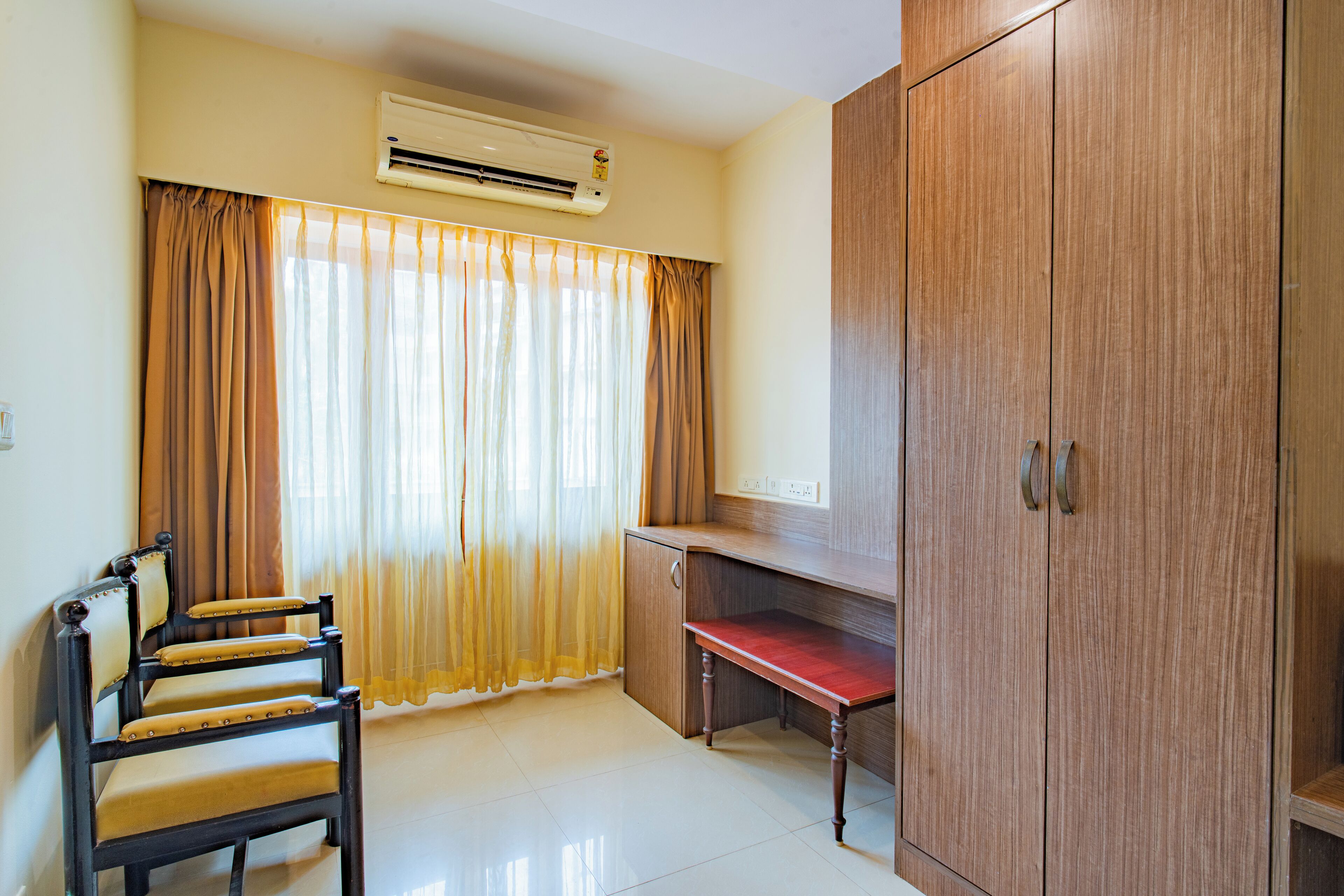 A room with a wooden wardrobe, a desk, a chair, and a window with curtains.