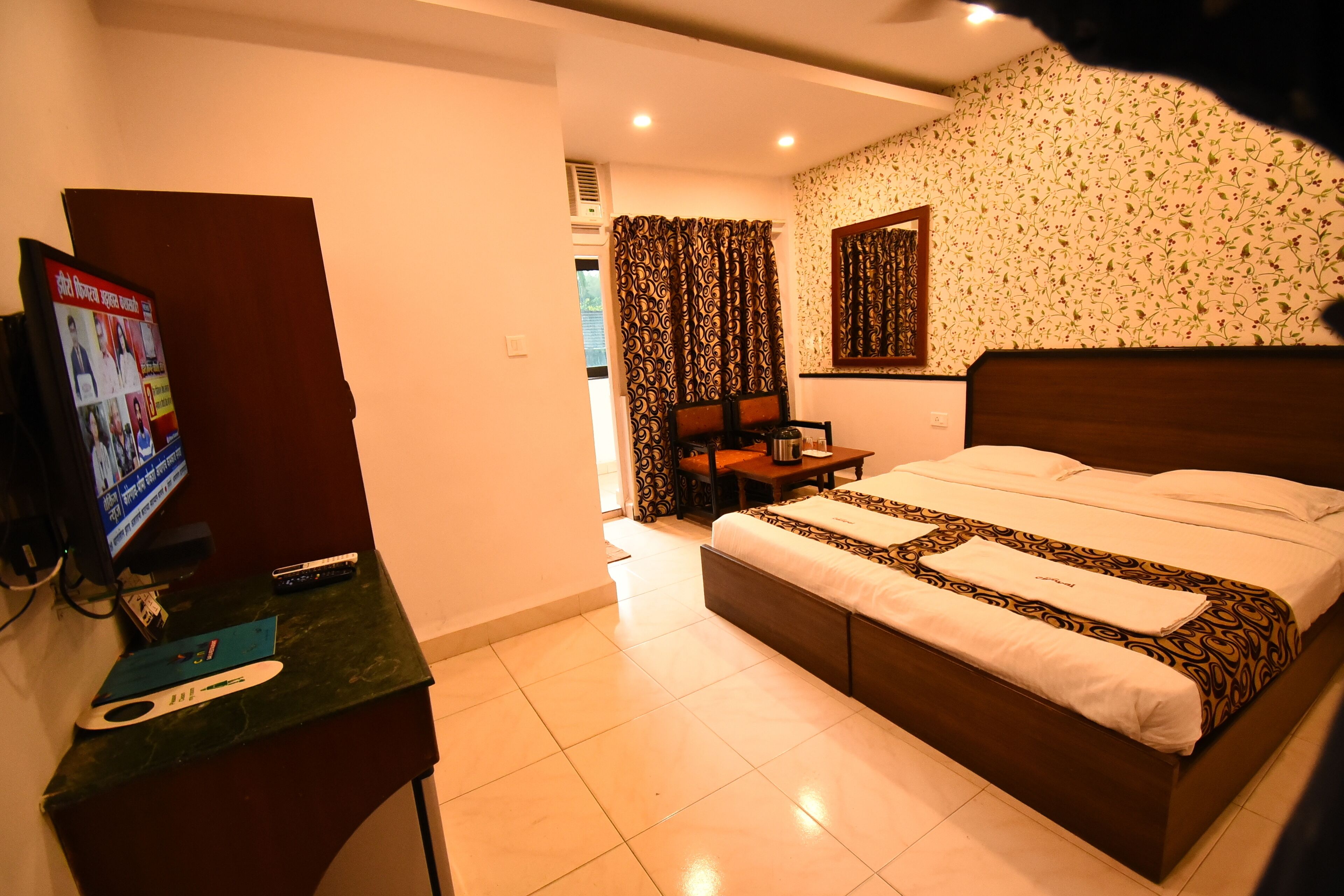 A hotel room with a bed, a TV on a stand, a desk with a remote control, and a patterned wall.