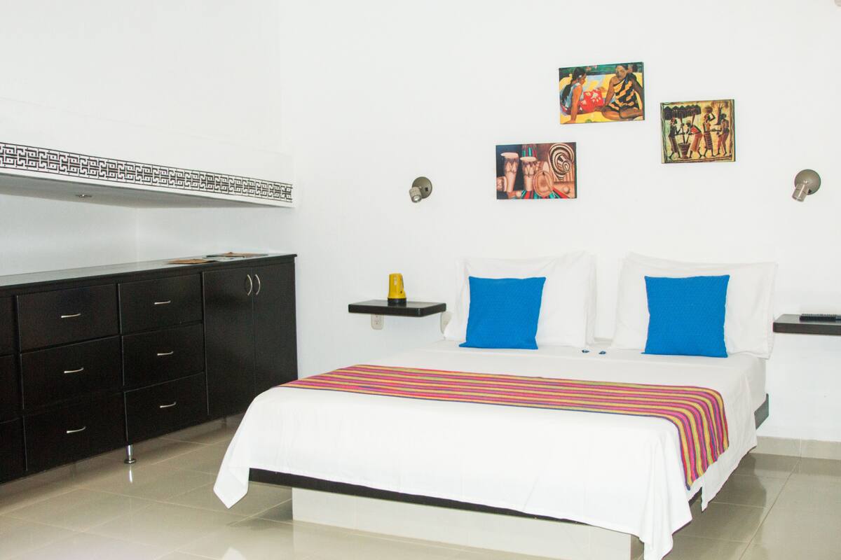 Una habitación de hotel con una cama, un armario negro, un estante con una botella amarilla y una obra de arte en la pared.