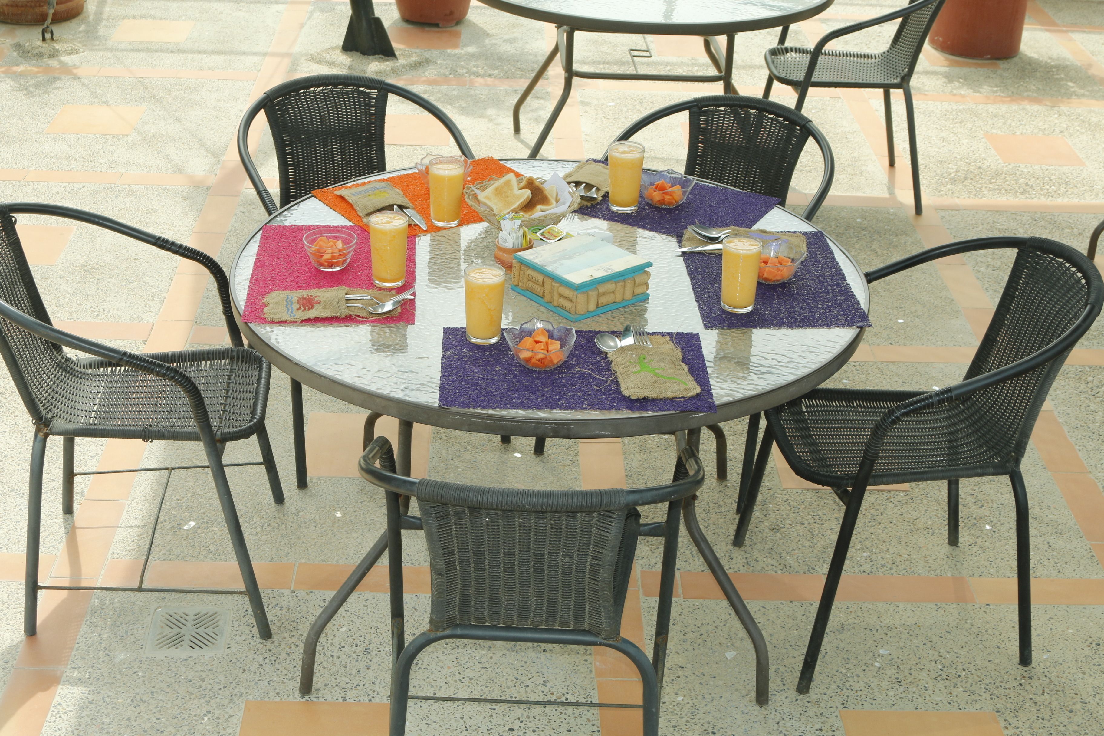 A round table with a glass top surrounded by black wicker chairs. The table is set with a purple placemat, a basket of bread, and several glasses of orange juice.
