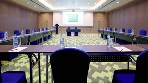 Un salón de conferencias con una larga mesa, sillas azules, botellas de agua y una pantalla para presentaciones.