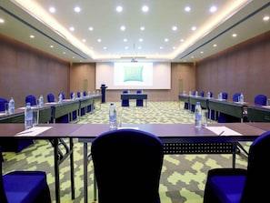 Un salón de conferencias con una larga mesa, sillas azules, botellas de agua y una pantalla para presentaciones.