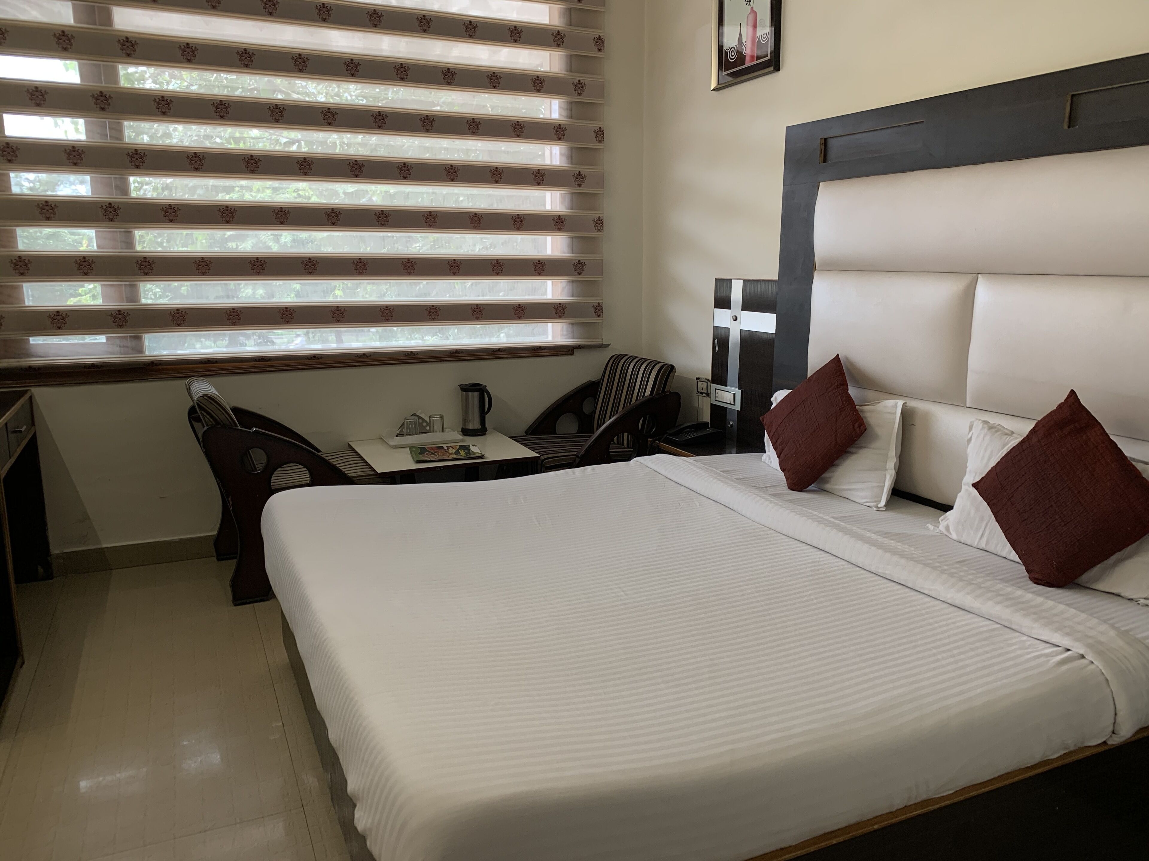 Habitación de hotel con una cama grande, un escritorio con silla, una cafetera y una ventana con persianas.