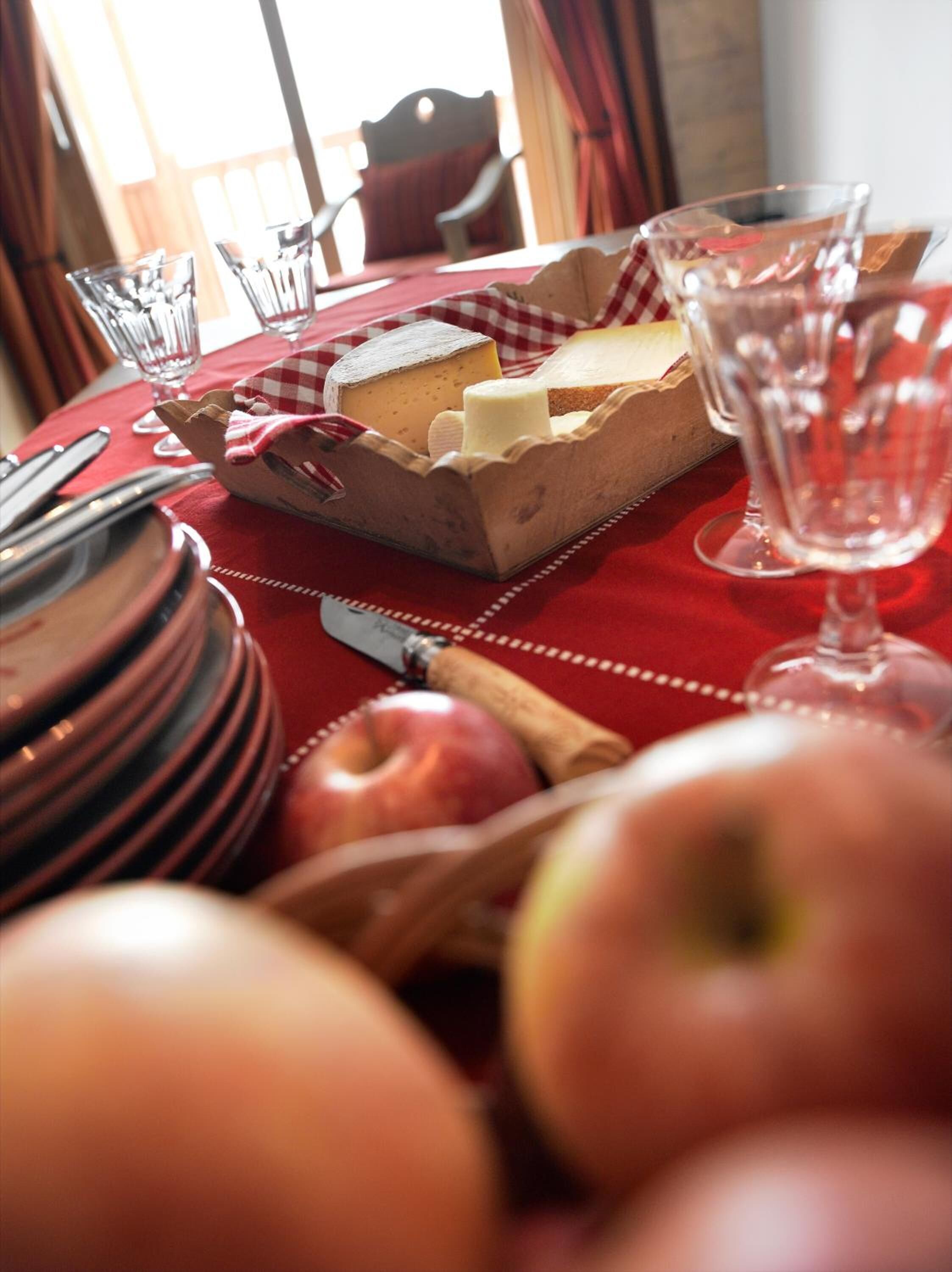 Une table à manger recouverte d’une nappe rouge, avec une planche à fromages, des pommes et un verre à vin.