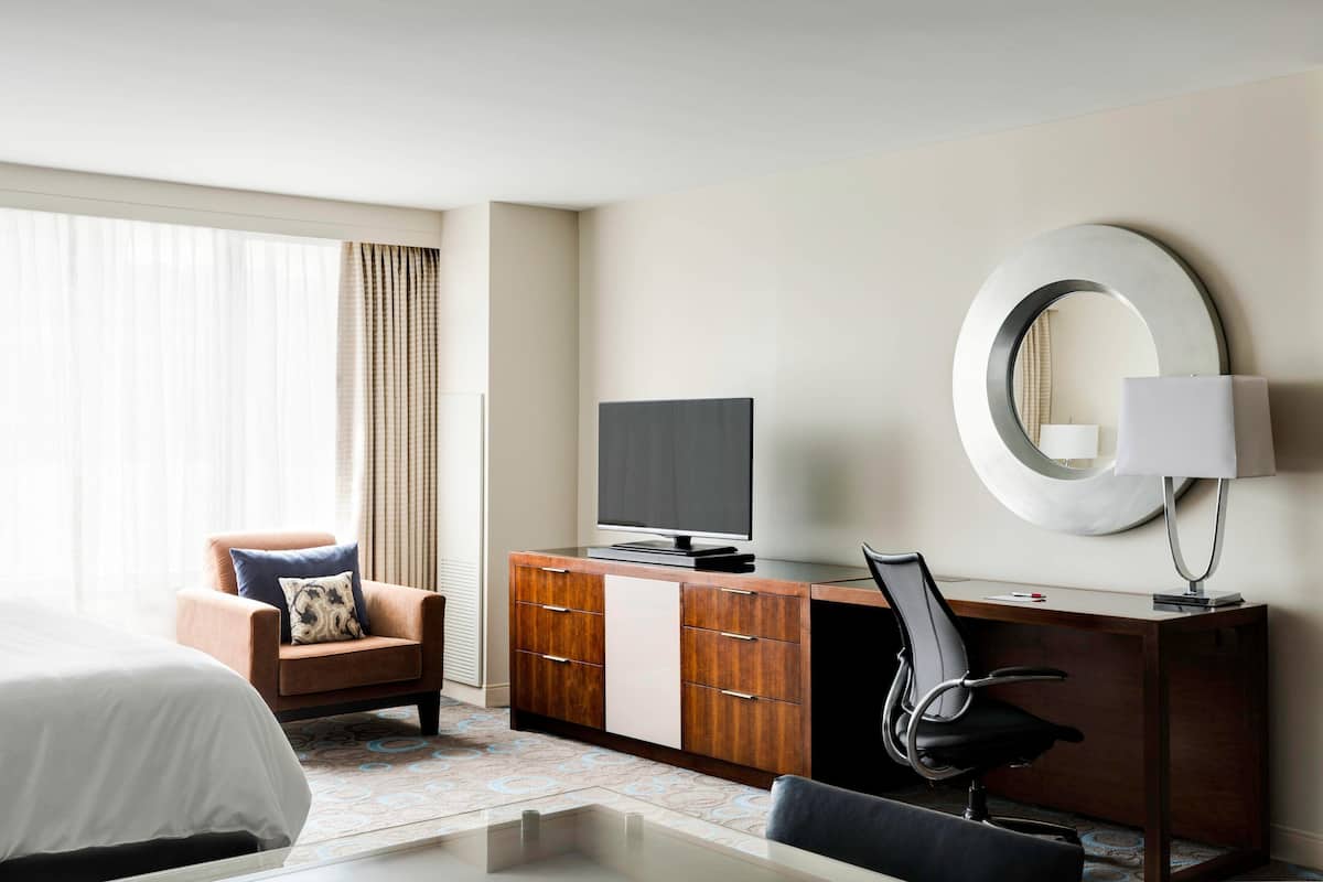 A hotel room with a bed, a desk with a TV, a chair, a lamp, and a large mirror on the wall.