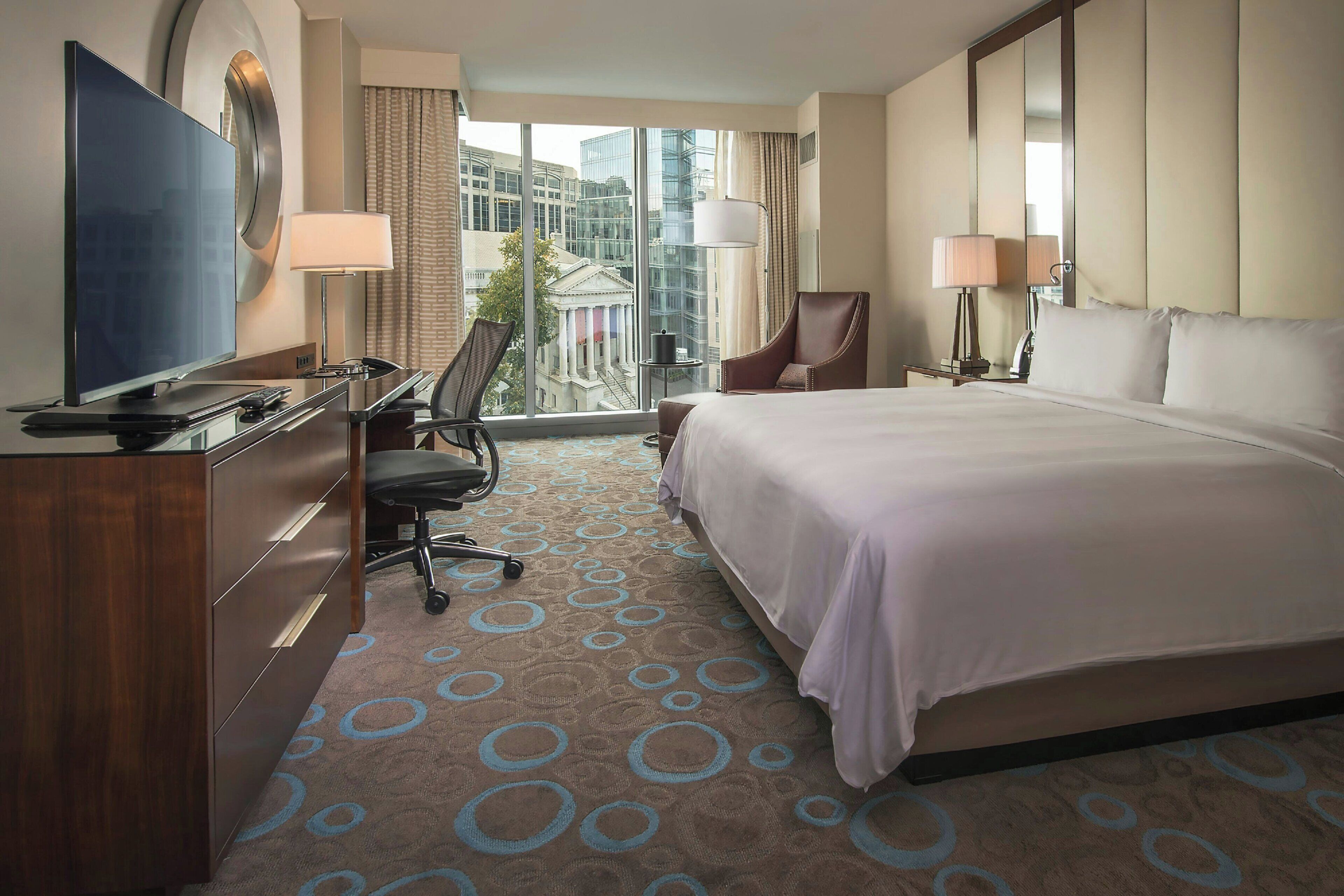 A hotel room with a large bed, a desk with a TV, a chair, and a view of a building through the window.