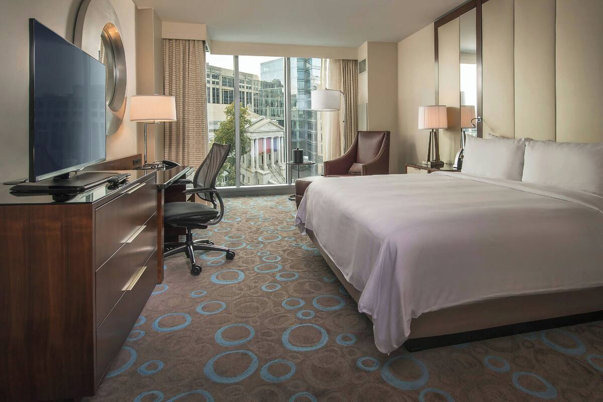 A hotel room with a large bed, a desk with a TV, a chair, and a view of a building through the window.