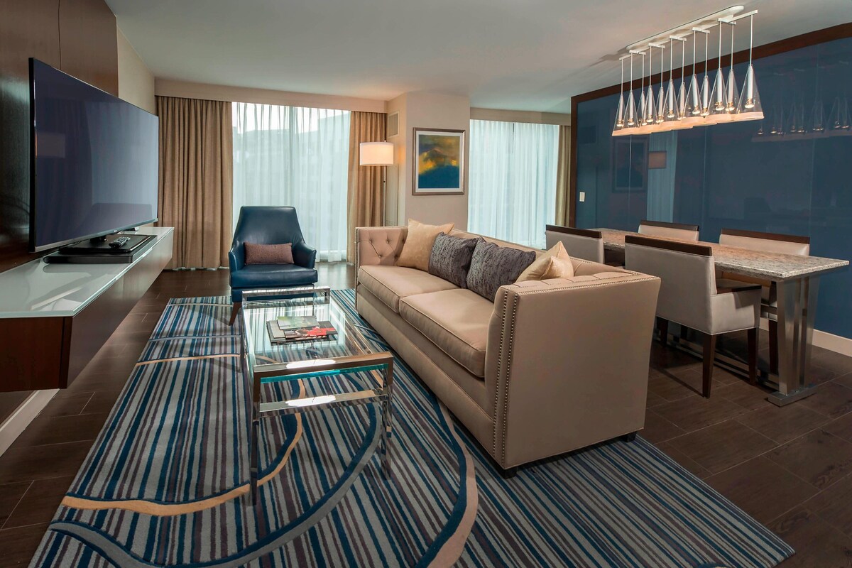 A modern hotel room with a large flat-screen TV, a sofa, a dining table, and a patterned rug.