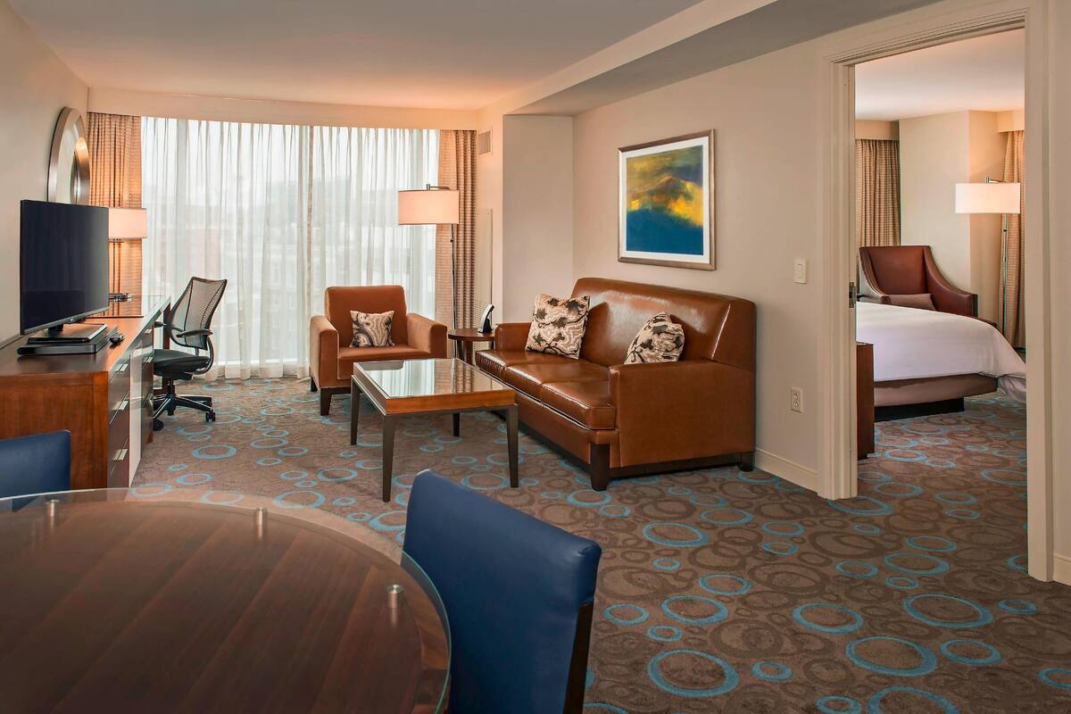 A hotel room with a brown leather sofa, a glass coffee table, a desk with a computer, a TV, and a bed with a white comforter.
