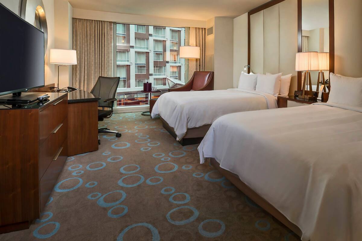 A hotel room with two beds, a desk with a TV, a chair, and a view of a building through large windows.