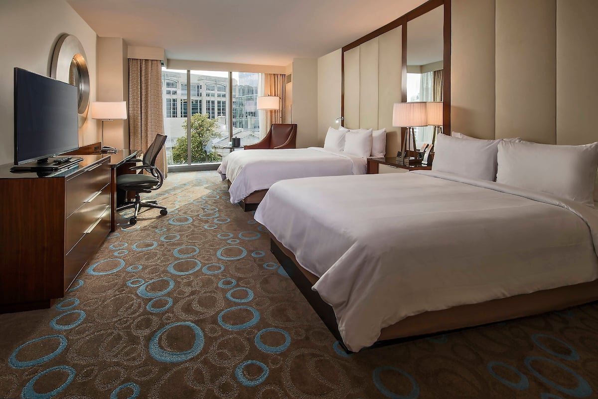 A hotel room with two beds, a desk with a TV, a chair, and a large window offering a city view.