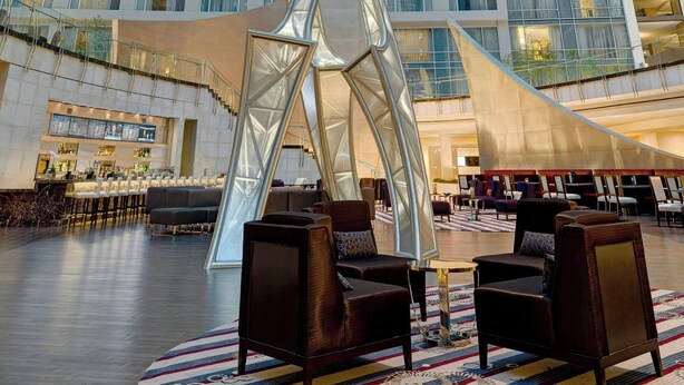 Un lobby moderno de hotel con un amplio techo de vidrio, una escultura abstracta en el centro y cómodas zonas de estar.
