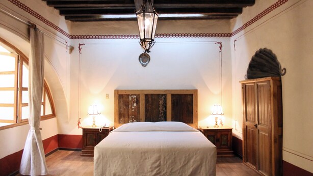 A bedroom with a large bed, wooden headboard, two bedside tables with lamps, a wardrobe, and a decorative ceiling light. - Hotel Casa de la Palma Travel (Puebla)