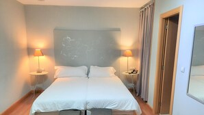 A double bed with white linens, two bedside tables with lamps, a headboard with a floral design, and a wooden floor. - Santana Hotel Restaurante (Palazuelos de Eresma)