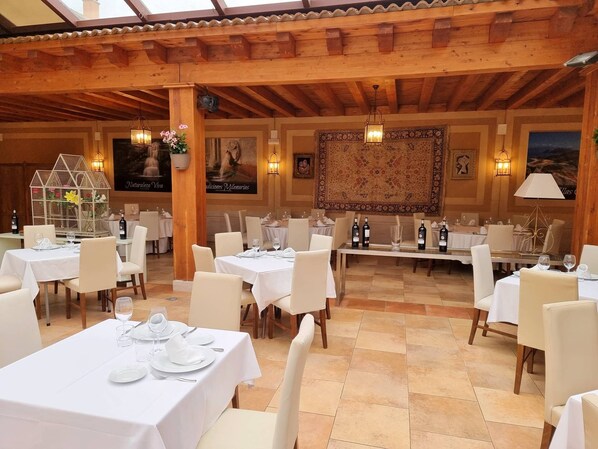 A restaurant with multiple tables set for dining, wooden beams on the ceiling, and a large decorative rug on the wall.