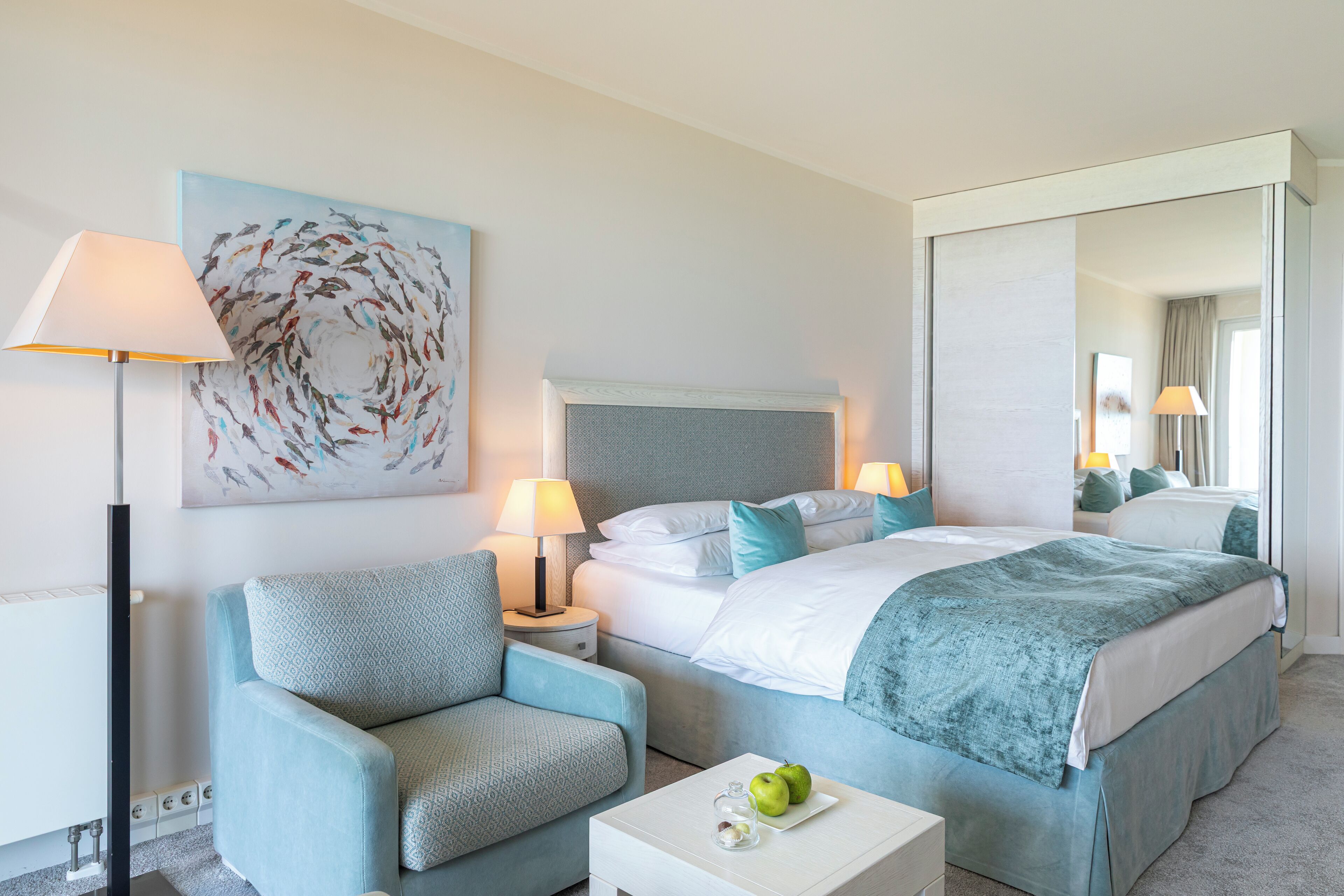 A hotel room with a bed, a chair, a bedside table, a lamp, and a painting on the wall.