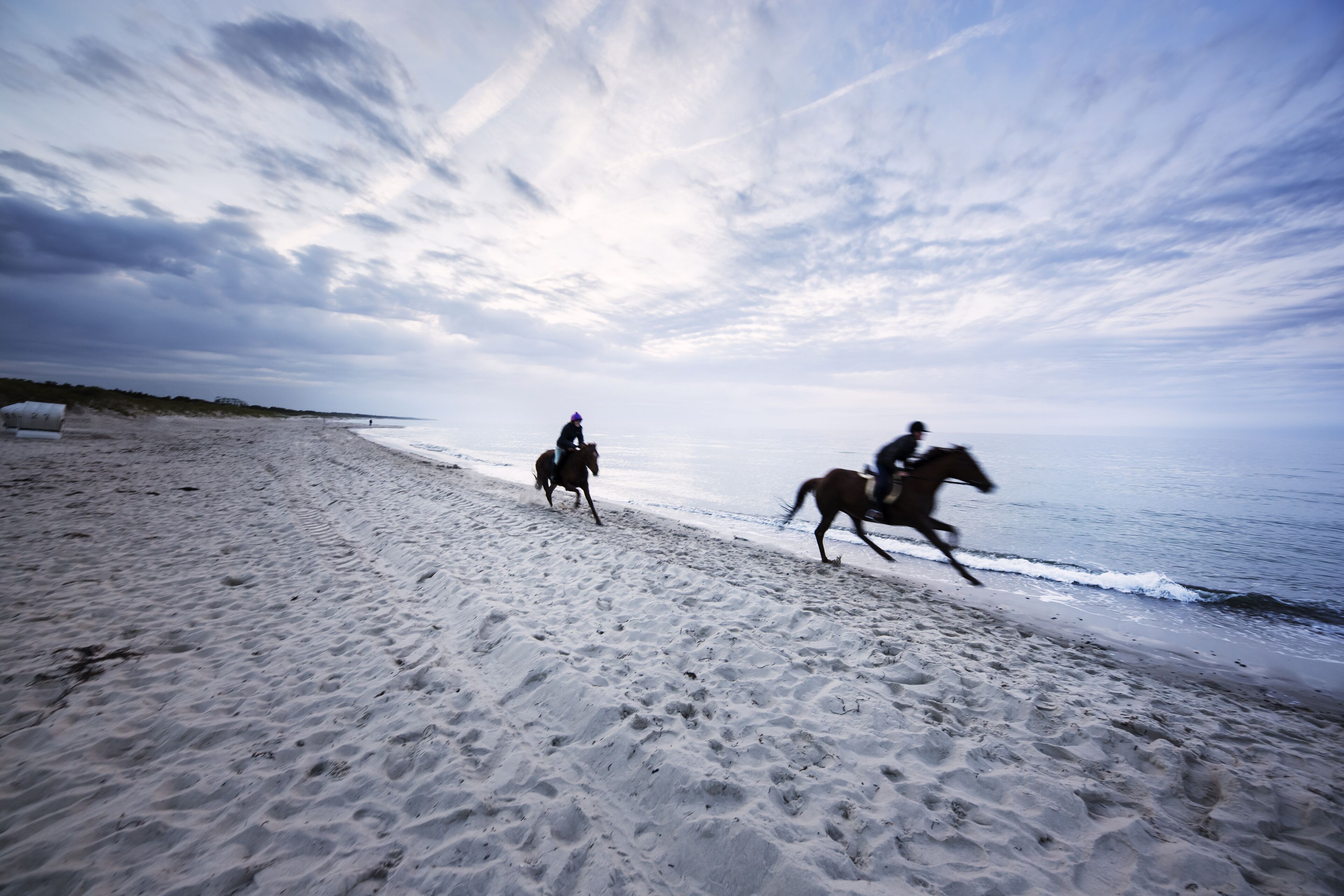 Una playa con dos personas montando a caballo por la orilla.