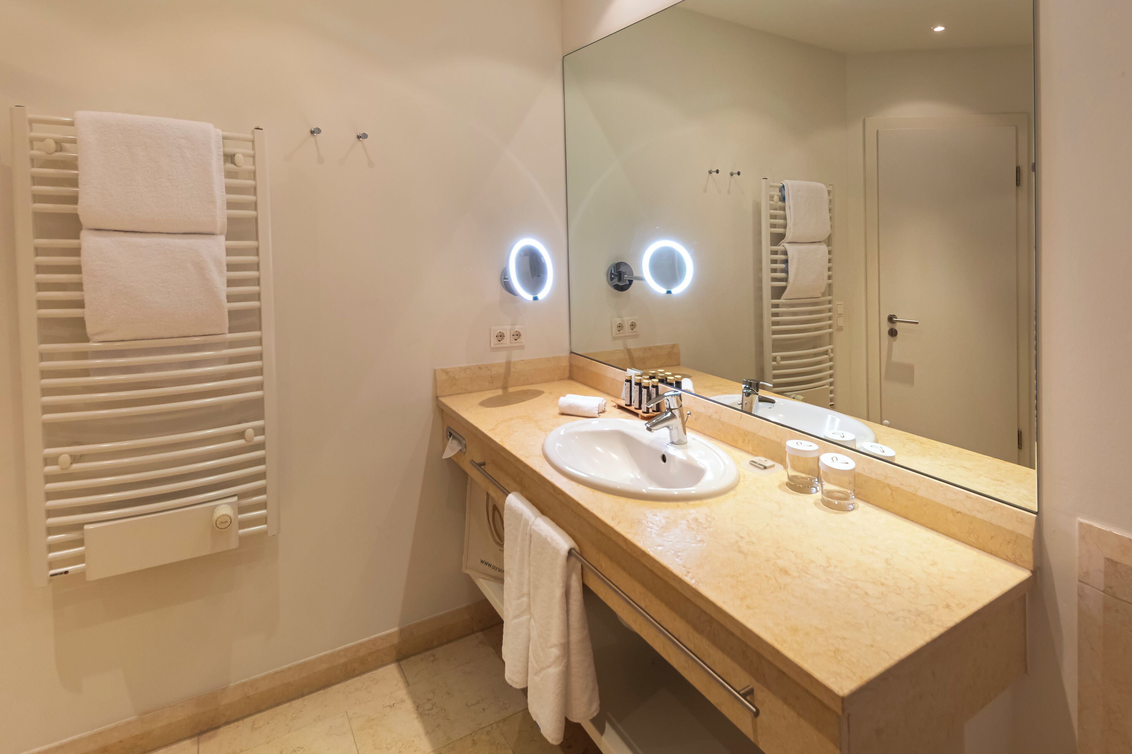 A modern bathroom with a beige countertop, a white sink, and a large mirror. There are two towel racks with white towels, and a wall-mounted heater.